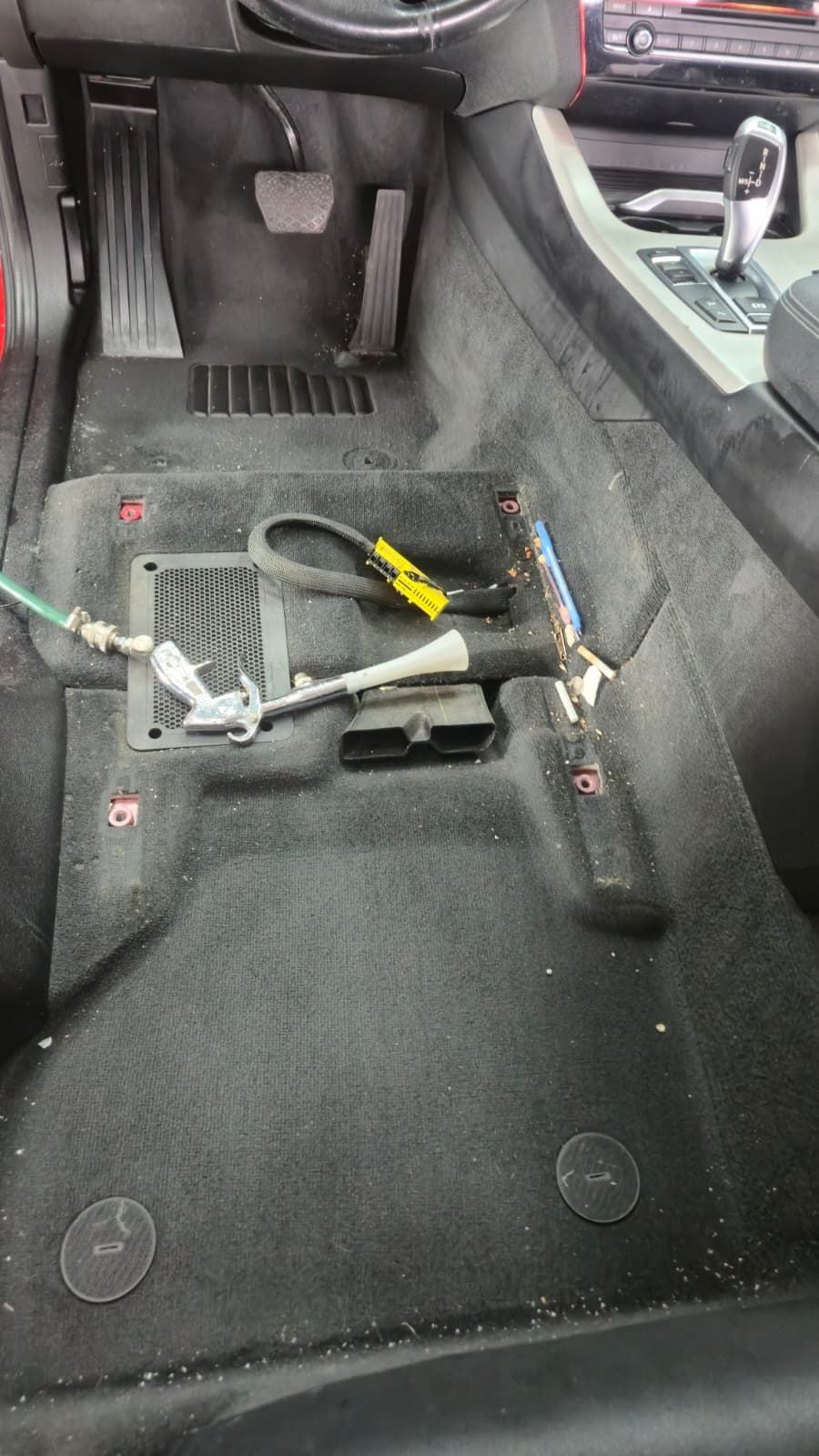 Podeaua din stânga față a unui BMW X3, cu scaunul șoferului demontat. Pe mochetă se văd firimituri, un pix și resturi adunate sub scaun, plus furtunul aparatului de extracție.